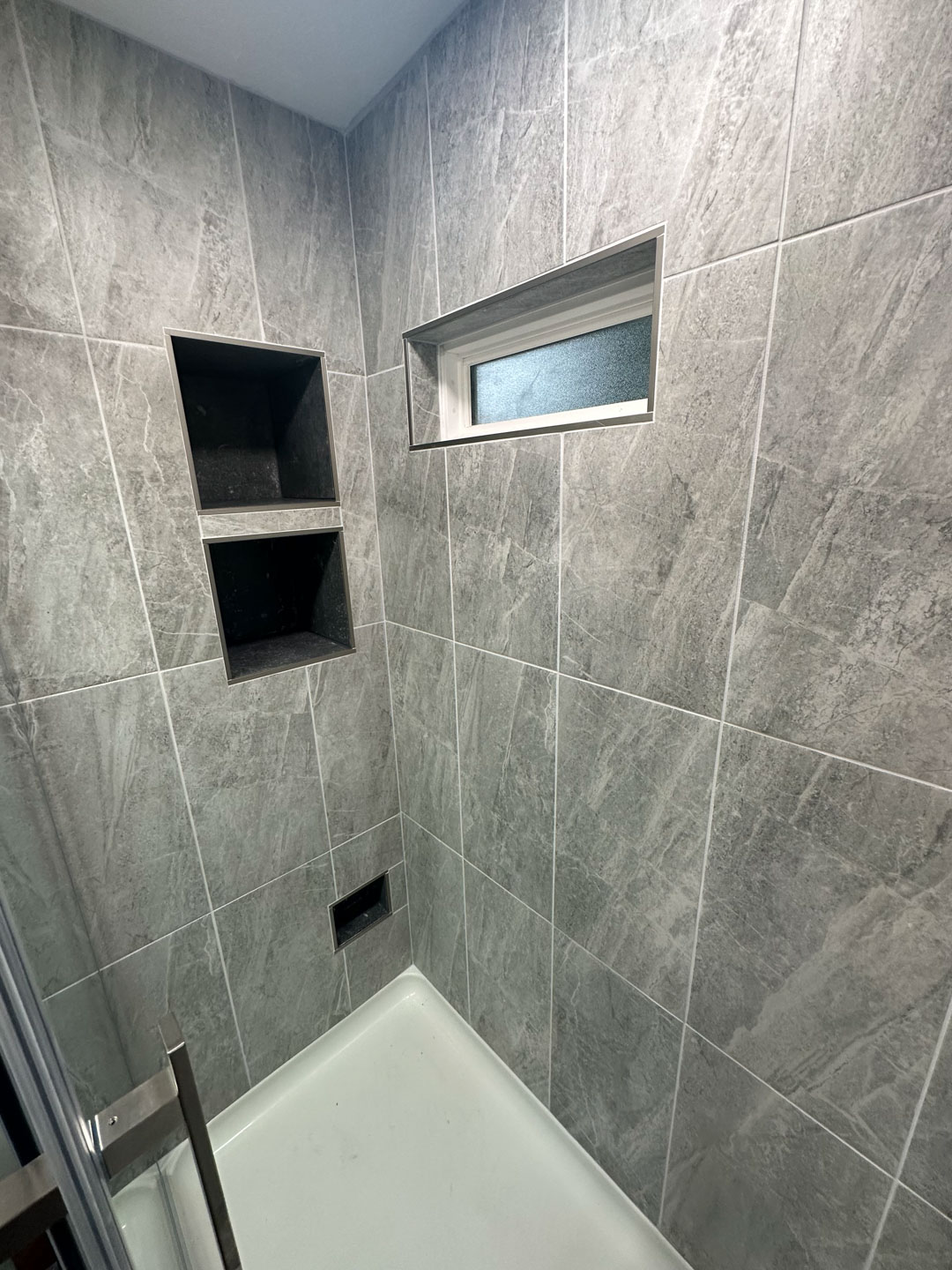 Corner of a shower with gray textured tiles, two built-in recessed shelves, a frosted horizontal window, and a white shower base.