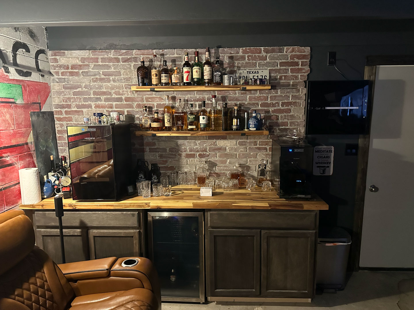 Home bar setup with wooden shelves holding various liquor bottles, glassware on a wooden countertop, two small refrigerators, and a brown leather recliner chair in front.