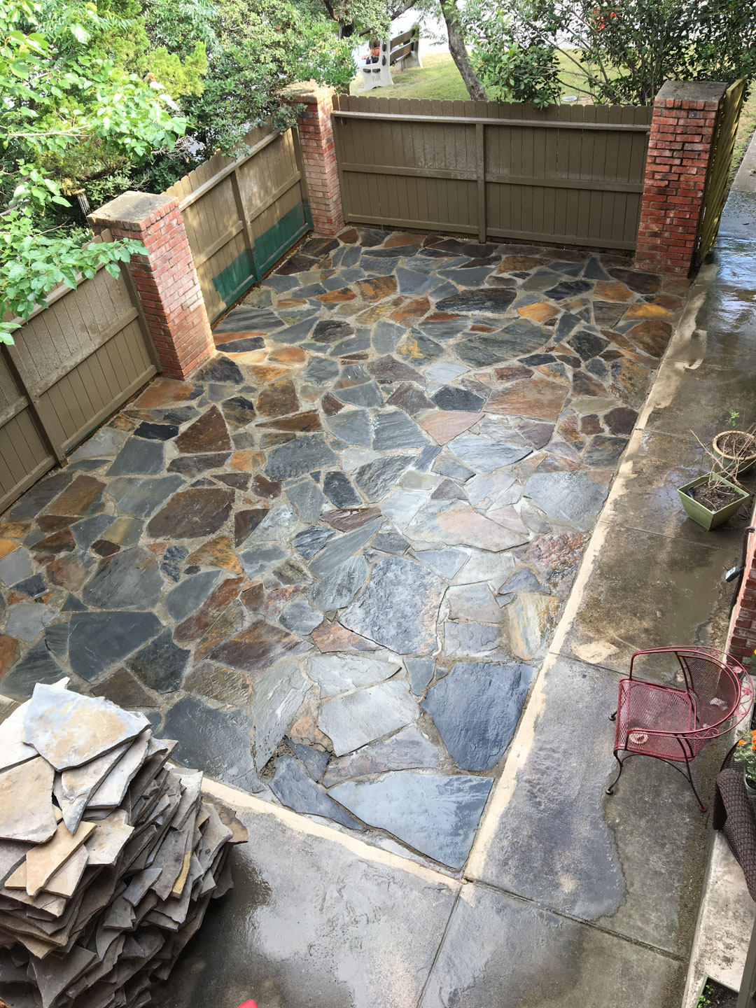Backyard patio with wet multicolored irregular stone tiles bordered by concrete, stacked stones on the left, and a red metal chair on the right.