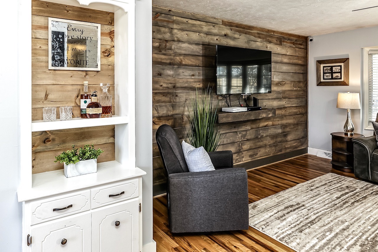 Cozy living room with wooden plank accent wall, mounted flat-screen TV, gray armchair, and white shelf with liquor bottles and glasses.