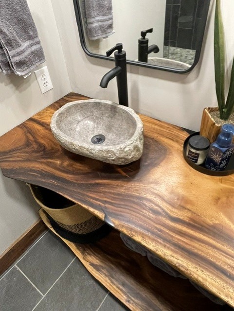 Natural wood bathroom vanity with a stone vessel sink, black faucet, gray towels, and decorative items on the countertop.