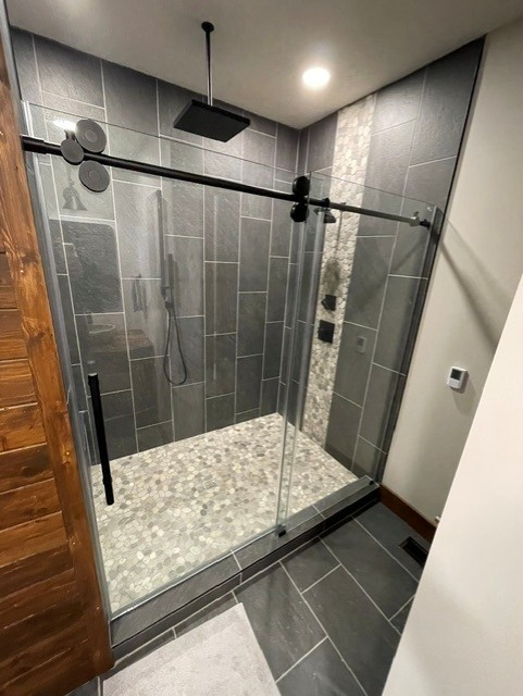 Modern walk-in shower with dark gray tile walls, pebble stone floor, glass sliding door, and black rain showerhead.