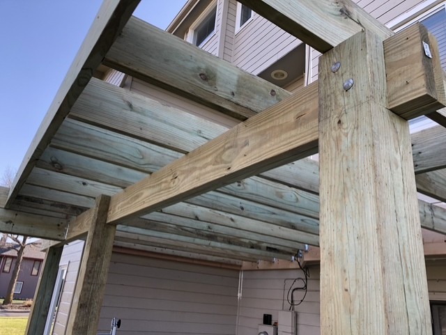 Close-up view of the wooden frame structure of a pergola attached to the side of a house.
